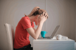 woman hunched over a laptop with hands clasped over her head, appearing stressed