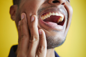 A close-up of a man holding his cheek with his mouth open, appearing to experience tooth or jaw pain against a yellow background