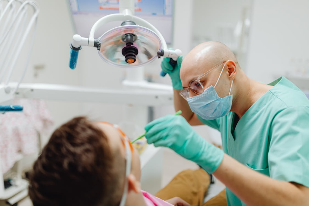 A dentist in green scrubs is shown treating a patient under dental light in a clinic